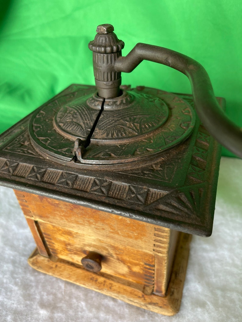 Op de afbeelding: Antiek gietijzeren koffiemolen met een houten basis en een lade. De molen heeft een decoratief ontwerp en een kruk.