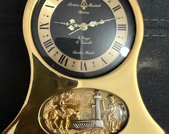 Puede incluir: Un reloj despertador musical de color dorado con una esfera negra y marcas de números romanos. La esfera del reloj dice "Arnex Musical Alarm 8 Days 15 Jewels Swiss Made". El reloj tiene una escena decorativa de un hombre a caballo y una mujer junto a una fuente.