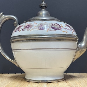 Könnte beinhalten: Eine weiße Porzellan-Teekanne mit einem silbernen Deckel und Henkel. Die Teekanne ist mit einem floralen Muster in Violett- und Brauntönen verziert.