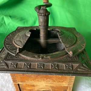 Op de afbeelding: Antiek gietijzeren koffiemolen met een houten lade en een houten kruk. De molen heeft een decoratief ontwerp op de bovenkant en zijkanten.