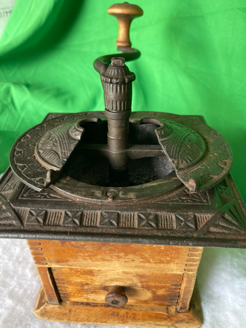 Op de afbeelding: Antiek gietijzeren koffiemolen met een houten lade en een houten kruk. De molen heeft een decoratief ontwerp op de bovenkant en zijkanten.
