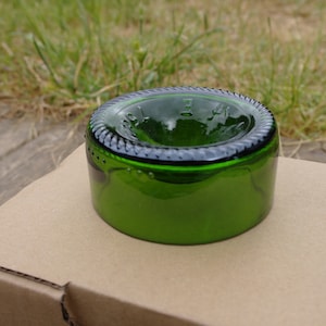 Peut inclure: Un dessus de bouteille en verre vert, coupé et poli pour créer un petit bol ou plat. Le dessus de la bouteille a un bord lisse et arrondi et une lèvre légèrement surélevée.