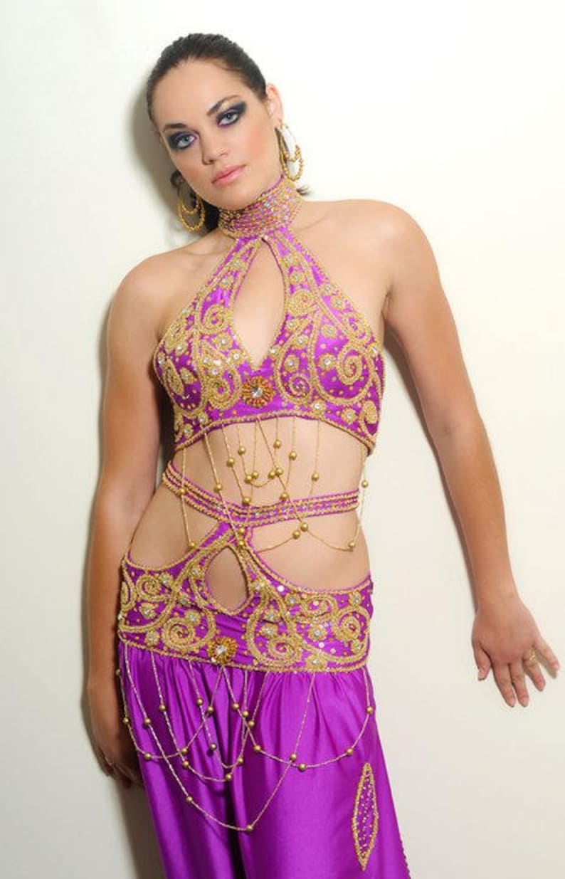 May include: A woman wearing a purple and gold belly dancing costume with intricate embroidery and beading. The costume includes a halter top, a belt, and pants.