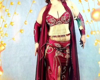 May include: A woman wearing a red and gold belly dancing costume with a long, flowing red skirt and a jeweled bra. The costume is decorated with gold sequins and beads. The woman is standing in front of a background of twinkling lights.