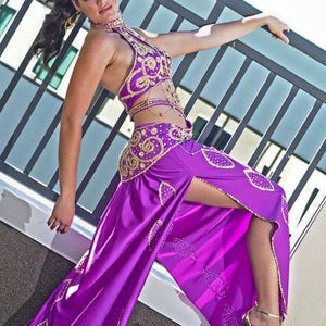 May include: A woman wearing a purple and gold belly dancing costume with a high slit skirt. She is posing with one leg raised and her hand on a railing.