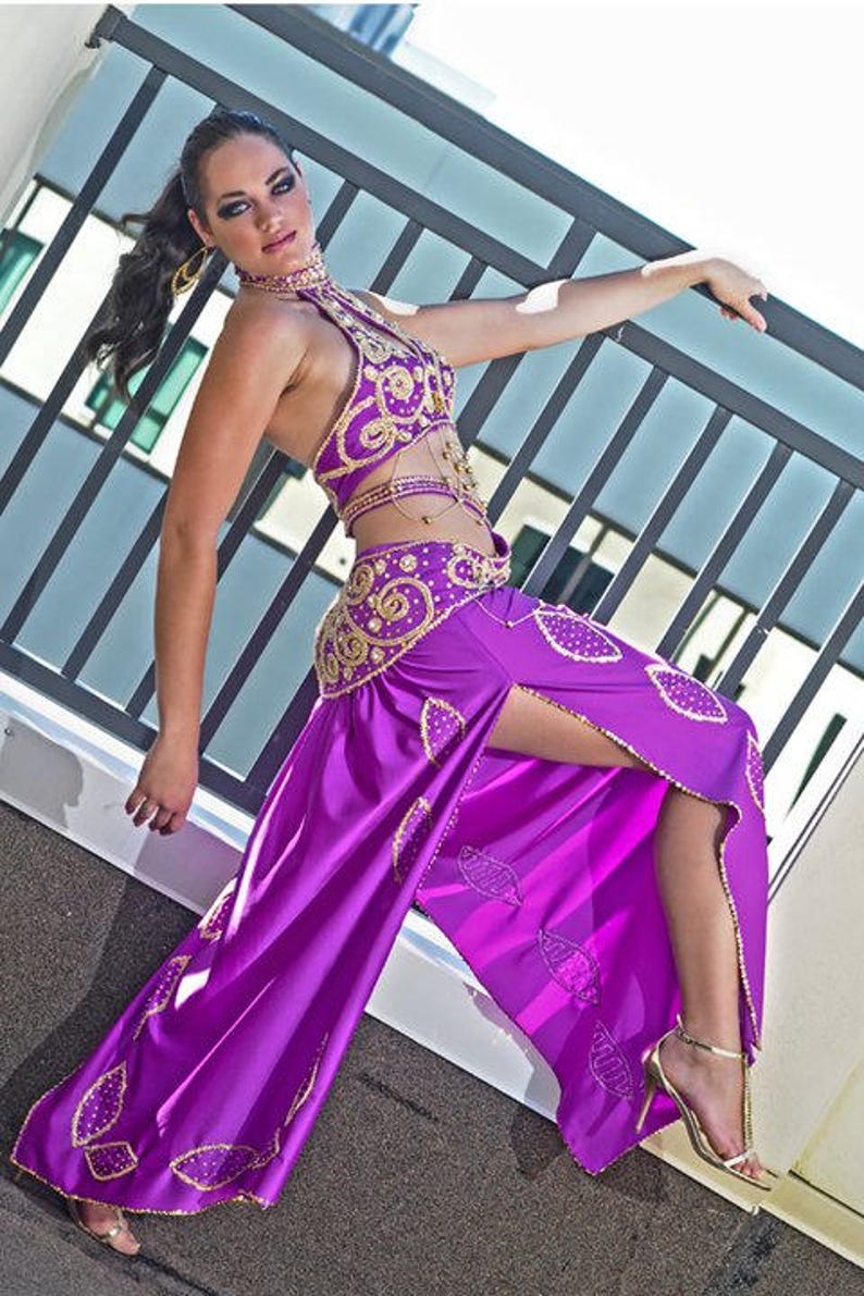 May include: A woman wearing a purple and gold belly dancing costume with a high slit skirt. She is posing with one leg raised and her hand on a railing.