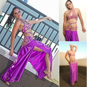 May include: A woman wearing a purple and gold belly dancing costume. The costume features a halter top, a skirt with a slit, and a belt with gold embellishments.