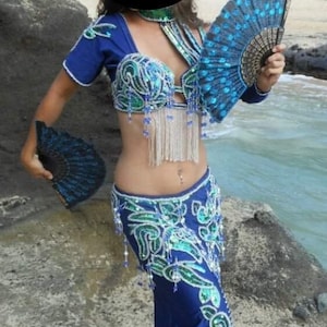 May include: A woman wearing a blue and green belly dancing costume with sequins and beads. She is holding two peacock feather fans.