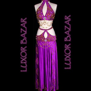 May include: A purple belly dancing costume with gold and silver embellishments. The costume includes a halter top, a belt, and a skirt. The skirt is long and flowing. The costume is decorated with sequins and beads. The text "Luxor Bazaar" is repeated on the image.