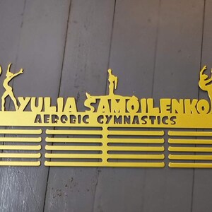May include: Gold metal medal display rack with 12 slots for medals. The rack features the name "YULIA SAMOILENKO" and the words "AEROBIC GYMNASTICS". The rack is decorated with three gymnast silhouettes.