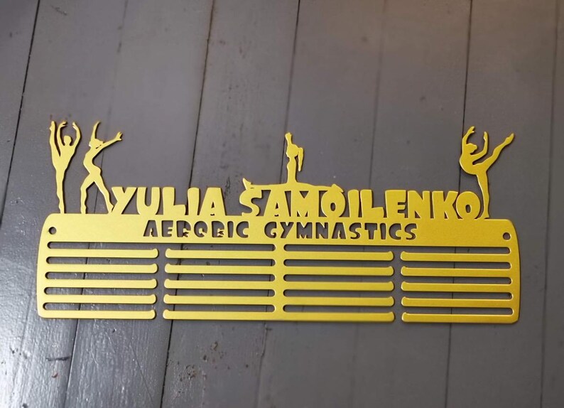 May include: Gold metal medal display rack with 12 slots for medals. The rack features the name "YULIA SAMOILENKO" and the words "AEROBIC GYMNASTICS". The rack is decorated with three gymnast silhouettes.