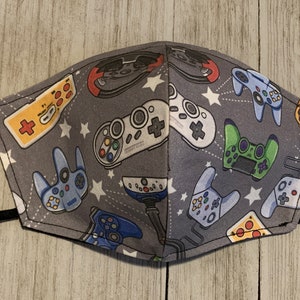 May include: Gray face mask featuring a pattern of colorful video game controllers and white stars. The controllers are in various colors, including red, yellow, green, and blue. Black elastic ear loops are attached.