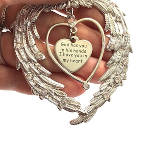 May include: Silver angel wing ornament with a heart-shaped charm that reads "God has you in his hands I have you in my heart".