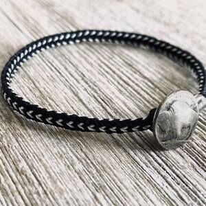 May include: A black and white braided bracelet with a silver coin charm. The coin has a raised design of a buffalo.