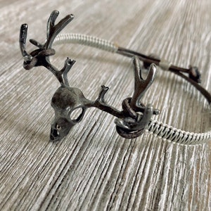 May include: A silver deer skull charm bracelet with a brown leather cord and a silver wire wrap.