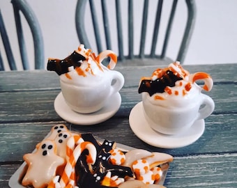 May include: Two miniature white coffee mugs with whipped cream, candy canes, and chocolate bats on top. The mugs are on white saucers. A tray of Halloween-themed cookies with orange and black icing, candy canes, and ghost shapes.