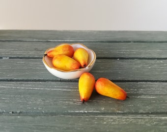 May include: A white bowl with a gold rim filled with six miniature yellow and orange pears. Two pears are sitting outside the bowl on a gray wooden surface.
