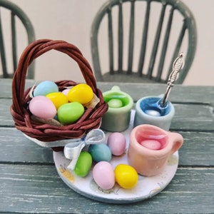 Puede incluir: Una cesta de mimbre en miniatura llena de huevos de Pascua de colores pastel, sentada sobre un plato blanco con más huevos y tres tazas en miniatura llenas de agua coloreada.