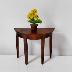 Pode incluir: Uma mesa de madeira semicircular com três pernas e uma planta em vaso com flores amarelas no topo.