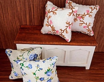 May include: Four miniature decorative pillows. Two pillows feature a white background with pink floral embroidery. The other two pillows have a white background with blue floral patterns. The pillows are arranged on a small white and brown wooden chest.