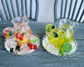 May include: Two glass pitchers filled with fruit-infused beverages, one with red fruit and the other with green fruit, are displayed on silver trays with glasses of the same beverages. The pitchers and glasses are decorated with fruit slices and mint leaves.