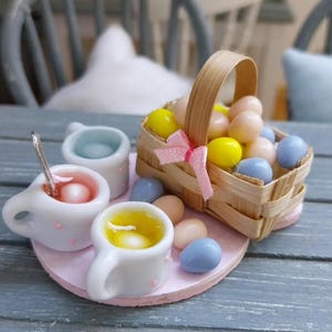 Puede incluir: Arreglo en miniatura con temática de Pascua, con una cesta tejida llena de huevos coloridos y un lazo rosa. Pequeñas tazas blancas con detalles rosas contienen líquidos y una cuchara. El arreglo está sobre una base circular rosa.