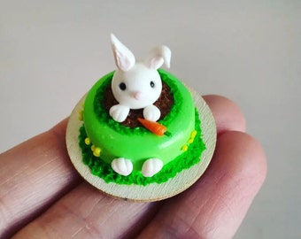 May include: A miniature cake decorated with green frosting and a white bunny peeking out of a brown hole. The bunny is holding a small orange carrot. The cake is on a round wooden base.