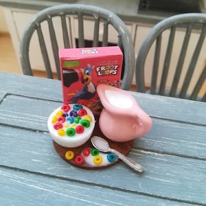 Puede incluir: Una escena de desayuno en miniatura con una jarra de crema rosa, un tazón de cereal Froot Loops de colores, una cuchara y una caja de cereal Froot Loops con un tucán.