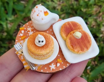 May include: Miniature Halloween breakfast scene featuring pancakes, a cup of coffee, and decorative elements. The pancakes are topped with a ghost-shaped dollop of cream and butter. The coffee cup is white with candy corn sprinkles. The scene is set on a small orange polka dot plate.