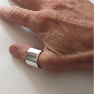 Peut inclure: Une bague de pouce en argent avec une bande large et plate. La bague est port&eacute;e sur le pouce d'une main.
