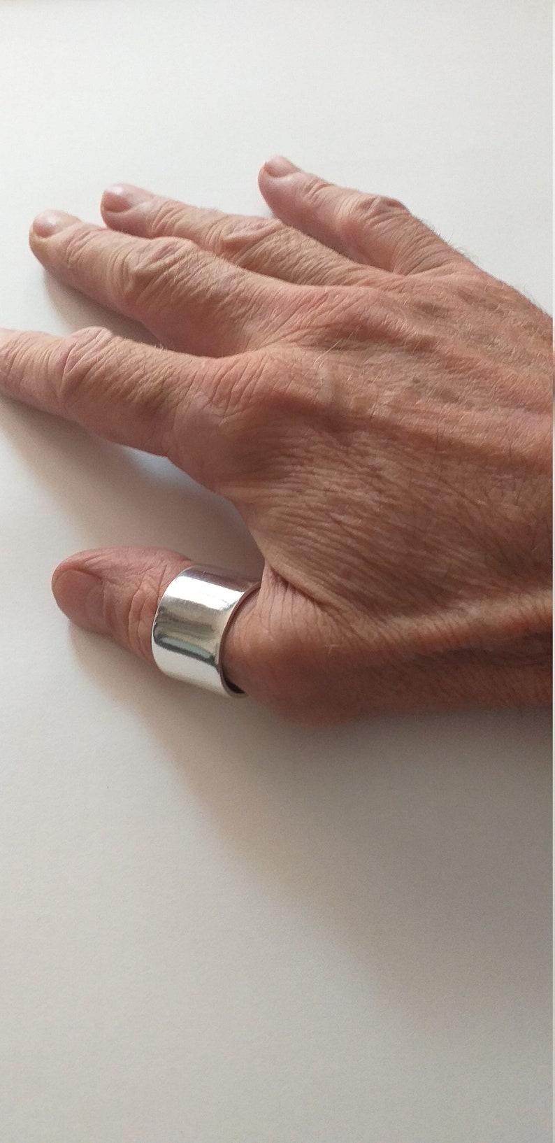 Peut inclure: Une bague de pouce en argent avec une bande large et plate. La bague est port&eacute;e sur le pouce d'une main.