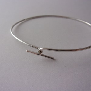 May include: A silver bangle bracelet with a simple, minimalist design. The bracelet has a t-bar clasp for easy opening and closing.