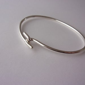 Könnte beinhalten: Ein silbernes Armreif-Armband mit einem schlichten, minimalistischen Design. Das Armband hat einen einzigartigen Verschluss, der aus einer Metallschachle gebildet wird.
