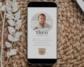 May include: A smartphone displaying a birthday invitation for a baby named Theo. The invitation features a photo of the baby and a cute cartoon bear. The text on the invitation reads "Theo is turning one!" and includes details about the party, including the date, time, and location. RSVP information is also included.