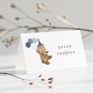 Może przedstawiać: Biała karta z napisem "pecan cookies" i akwarelową ilustracją brązowego misia w czapeczce urodzinowej i trzymającego niebieskie balony. Karta jest ustawiona pod kątem na białej powierzchni.
