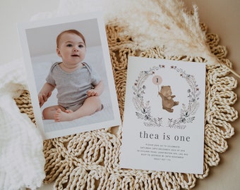 May include: A white card with a floral wreath and a brown bear holding a pink balloon with the number 1. The text "thea is one" is below the wreath. The card is on a woven straw mat with a white photo card of a baby girl sitting on a white surface.