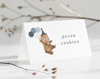 May include: A white card with the text "pecan cookies" and a watercolor illustration of a brown bear wearing a party hat and holding blue balloons. The card is angled on a white surface.