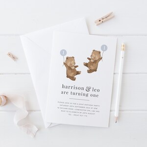 May include: A white invitation card featuring a brown bear illustration holding a blue balloon with the number one on it. The text on the card reads "harrison & leo are turning one". The invitation includes details for a joint birthday party.