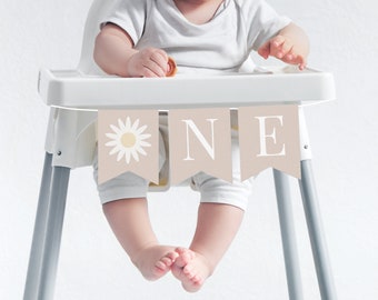 Op de afbeelding: Een baby die in een kinderstoel zit met een banner waarop "ONE" staat in witte letters op een beige achtergrond. De banner heeft een witte madeliefje met een gele kern aan de linkerkant.