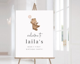 May include: Welcome to Laila's Bear-y First Birthday Party! A white sign with a brown bear holding a pink balloon. The bear is wearing a brown bow tie.