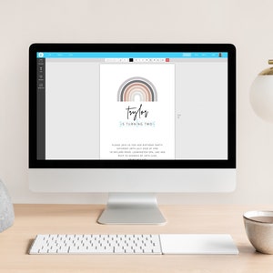 May include: A computer monitor displays a birthday invitation design. The invitation features a rainbow graphic in neutral tones above the name "Taylor" and the text "is turning two." The invitation is on a desk with a lamp and a coffee cup.