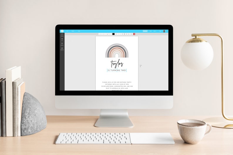 May include: A computer monitor displays a birthday invitation design. The invitation features a rainbow graphic in neutral tones above the name "Taylor" and the text "is turning two." The invitation is on a desk with a lamp and a coffee cup.