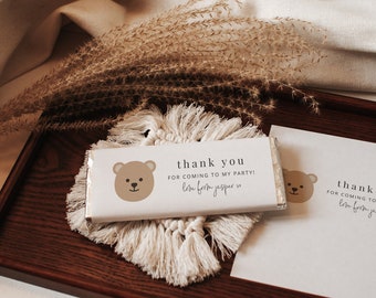 May include: Two white chocolate bar wrappers with a brown bear illustration and the text "thank you for coming to my party! love from jasper" on a wooden tray with a white woven mat.