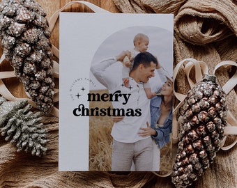 May include: A Christmas card with a photo of a family of three. The card is white with black text that says "The Williams Family + merry christmas". The photo shows a man holding a young child on his shoulders, while a woman stands next to him, smiling. The photo is set against a backdrop of tall grass.