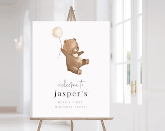 May include: A welcome sign for a birthday party featuring a cartoon brown bear holding a white balloon. The sign reads "Welcome to Jasper's Bear-y First Birthday Party!"