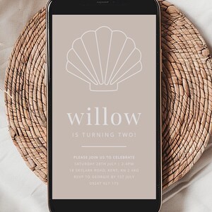 May include: A smartphone displays a digital invitation with a seashell graphic. The text reads "willow IS TURNING TWO!" and includes event details. The phone rests on a woven, circular placemat.