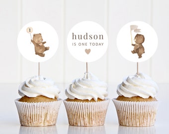 May include: Three cupcakes with brown bear themed toppers. The toppers are round and have a white background. The first topper has a brown bear holding a balloon with the number "1" on it. The second topper says "hudson is one today" with a small pink heart. The third topper has a brown bear holding a white flag.