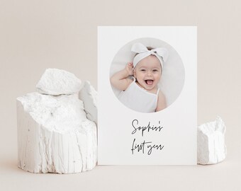 Puede incluir: Una tarjeta blanca con una foto circular de una niña bebé con una diadema blanca. El texto "Sophia's first year" está escrito en cursiva negra debajo de la foto.
