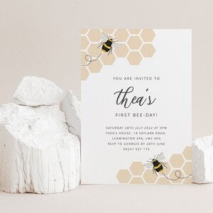 K&ouml;nnte beinhalten: Eine wei&szlig;e Karte mit einer bienenf&ouml;rmigen Einladung zu einer ersten Geburtstagsfeier. Die Karte hat ein Wabenmuster mit einer Biene auf jeder Seite. Der Text lautet "You are invited to Thea's First Bee-Day!" mit Datum, Uhrzeit und Ort der Party.