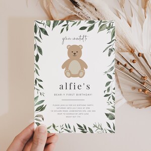 Puede incluir: Una tarjeta de invitaci&oacute;n blanca con un borde de hojas verdes. La tarjeta presenta un oso de peluche marr&oacute;n de dibujos animados y el texto "you're invited to alfie's BEAR-Y FIRST BIRTHDAY!" La invitaci&oacute;n incluye detalles para una fiesta de cumplea&ntilde;os, incluida la fecha, la hora, la ubicaci&oacute;n y la informaci&oacute;n de RSVP.